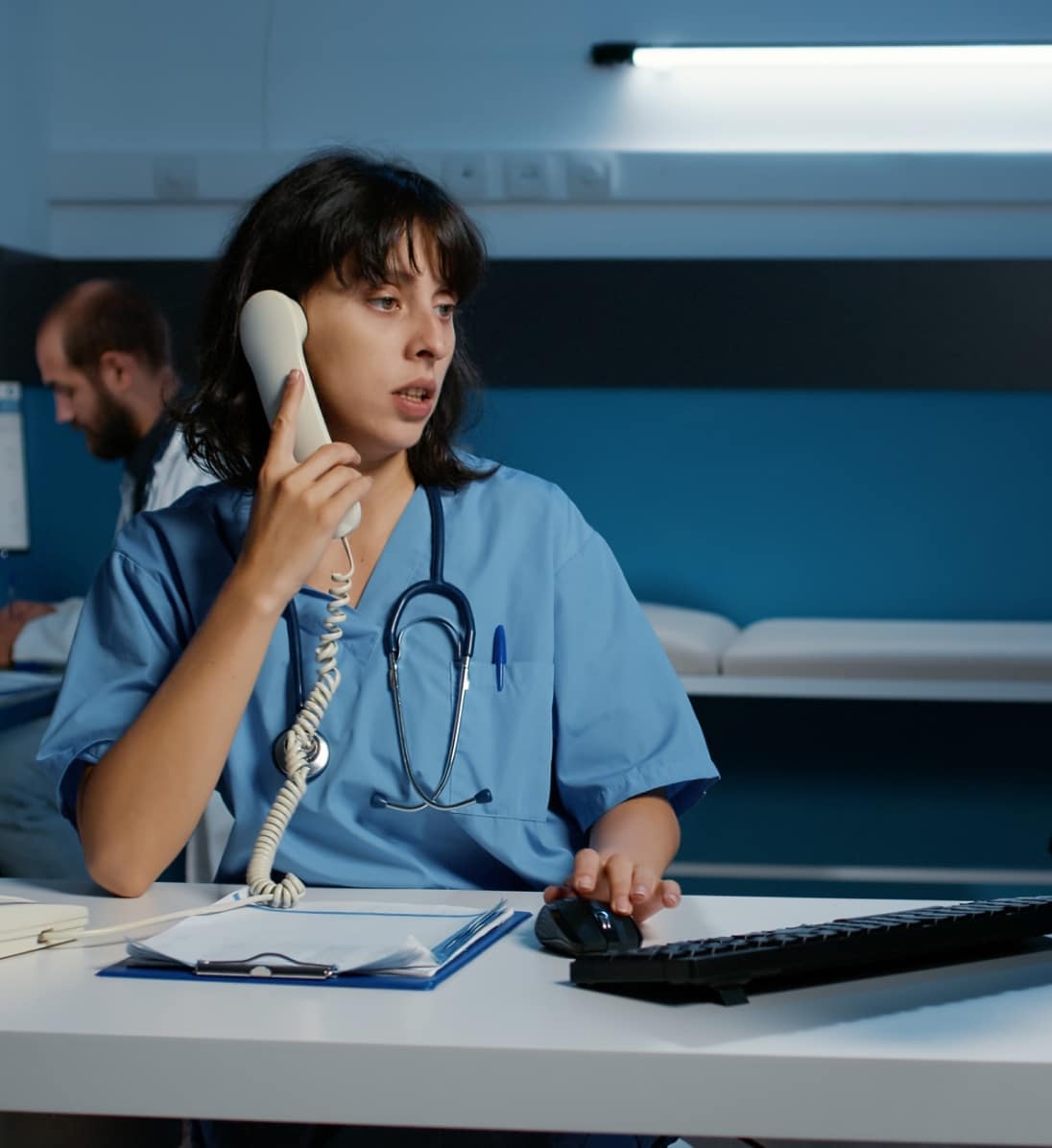 Medical provider standing at a desk, talking on a phone, while moving the mouse courser, looking over at computer screen.