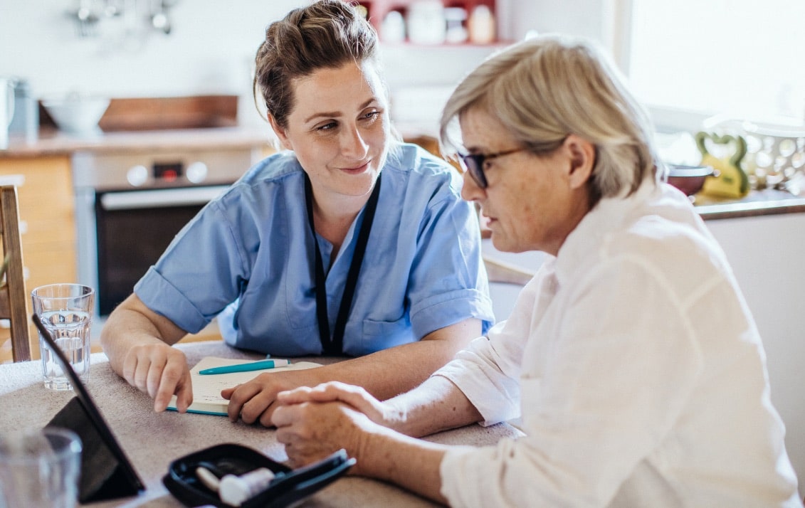 A nurse sits with a patient reviewing their healthcare plan on a tablet sitting on a table.