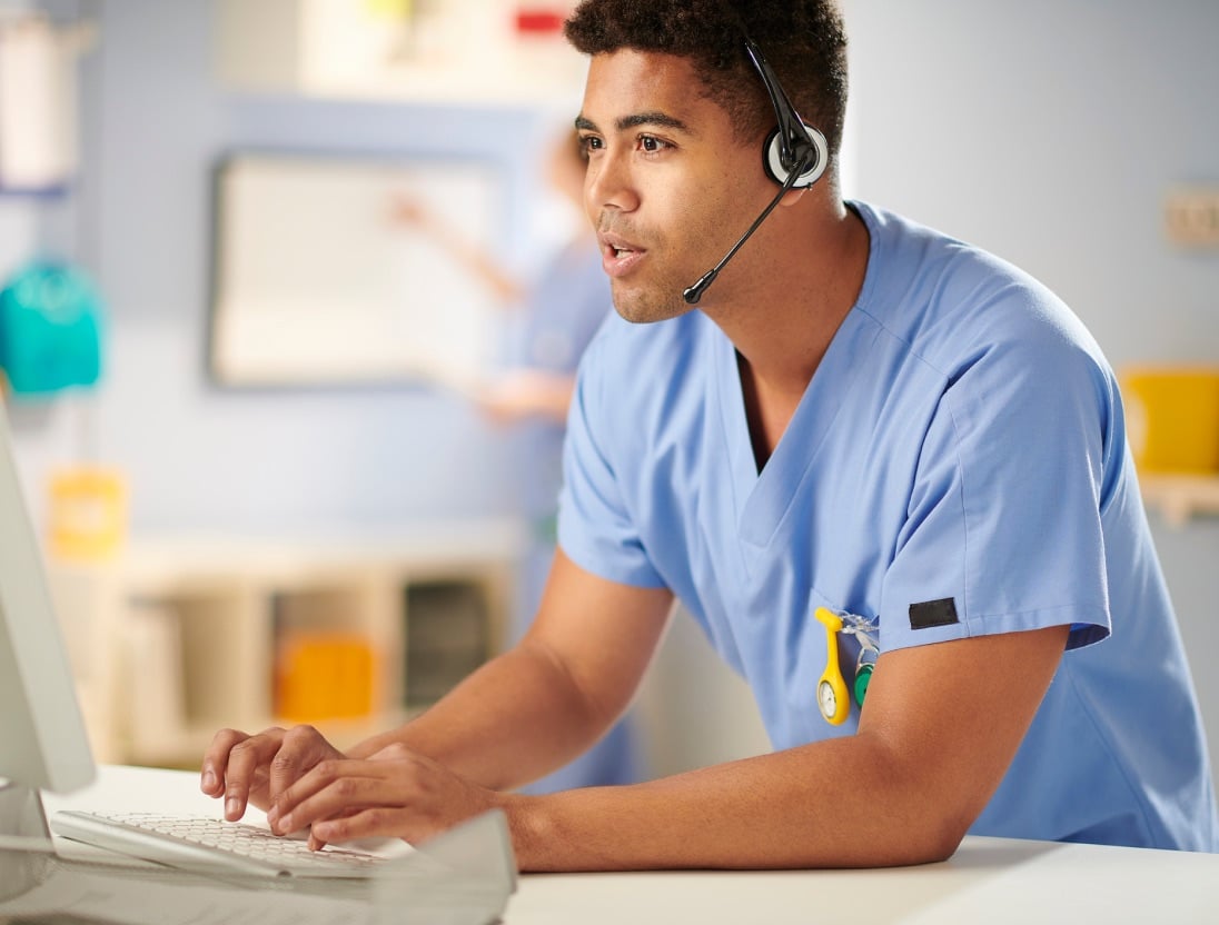 Healthcare professional wearing a headset types on a keyboard as they talk with a patient.