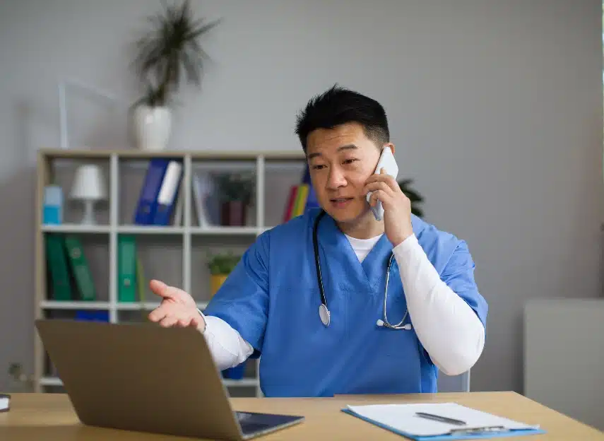 Medical provider talking on a cellphone while making a hand gesture at their laptop.