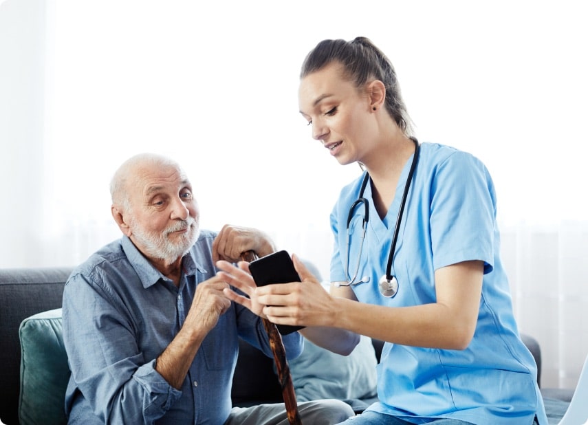 Elder man sits, leaning on his cane, while a nurse holding out a cellphone shows him something on the screen.