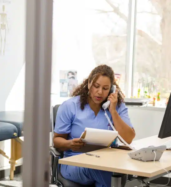 A nurse sitting at a desk, talking on a corded phone, while holding a clipboard and papers.
