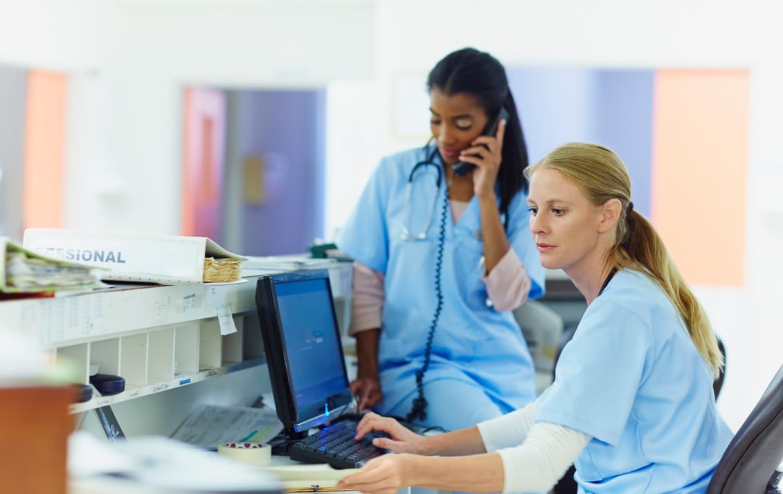 One nurse looks over patient charts as another sits in the background on a corded phone.