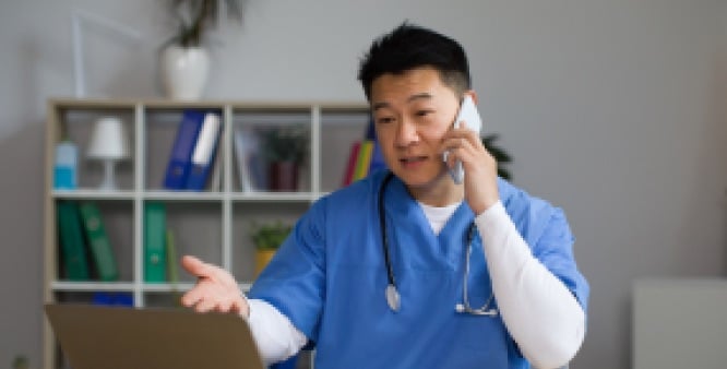 Healthcare professional standing, talking on a phone, with their other hand extended out towards an open laptop.