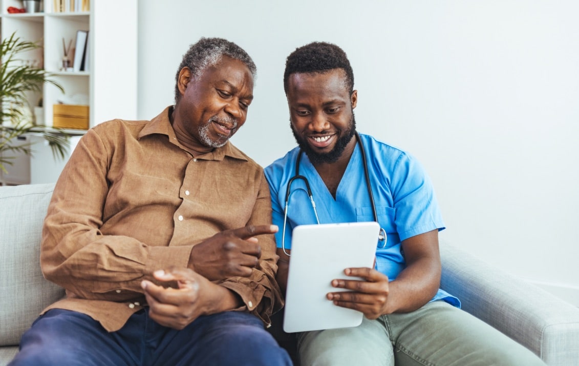 An elderly man and a nurse sit on a couch looking at a tablet.