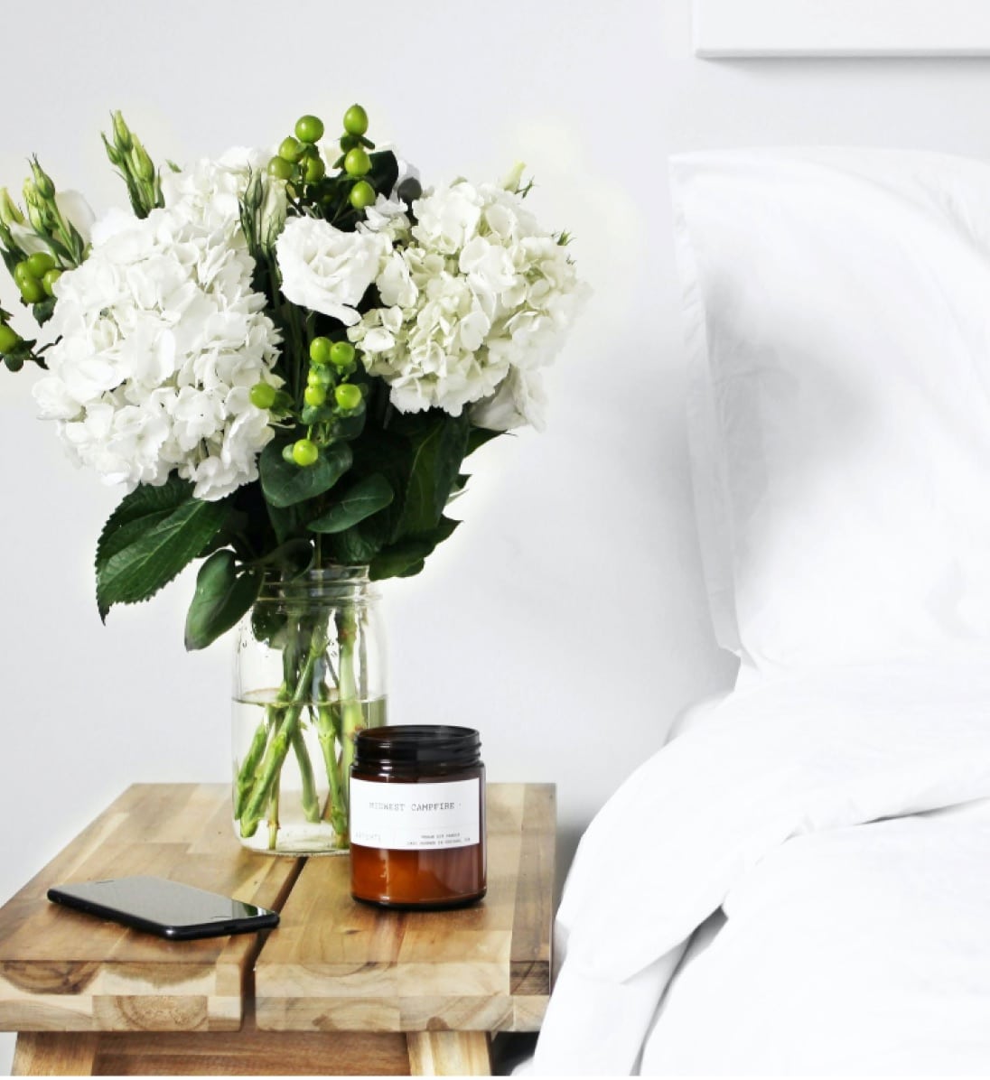Vase sits on a nightstand with white flowers next to a bed.