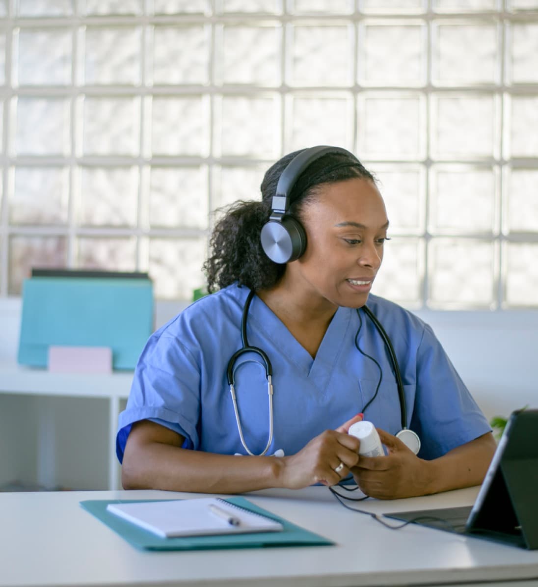 Nurse wearing a headset with a medicine bottle in hand, while virtually talking with a patient.