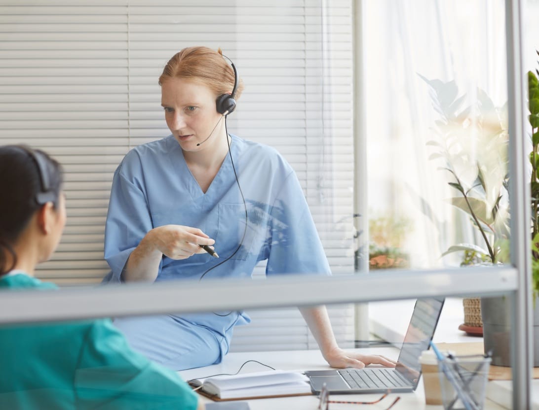 Two medical providers wearing headsets, one sitting on a desk, looking at each other.