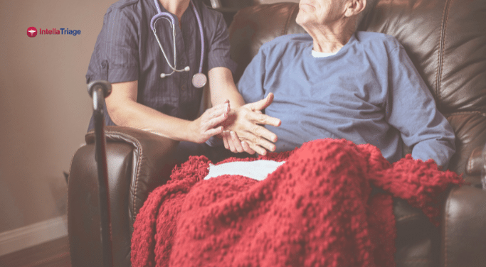 A nurse kneels beside an elderly man sitting in an armchair with a red blanket, offering comfort and compassionate care.